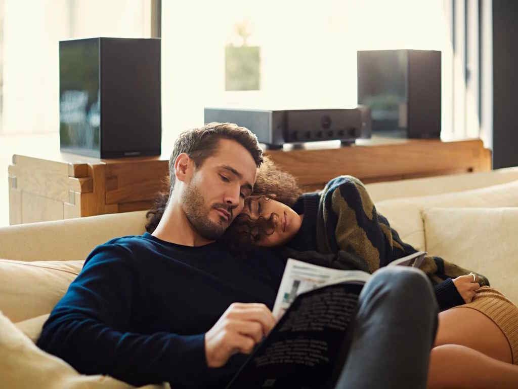 Two people relaxing on a beige couch, one reading while the other leans on their shoulder, with speakers and an audio receiver behind them.