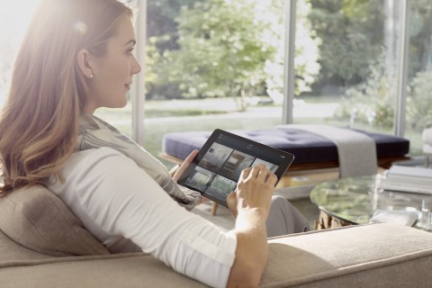 A woman uses the Savant application on a tablet to monitor security cameras while relaxing on the living room couch.