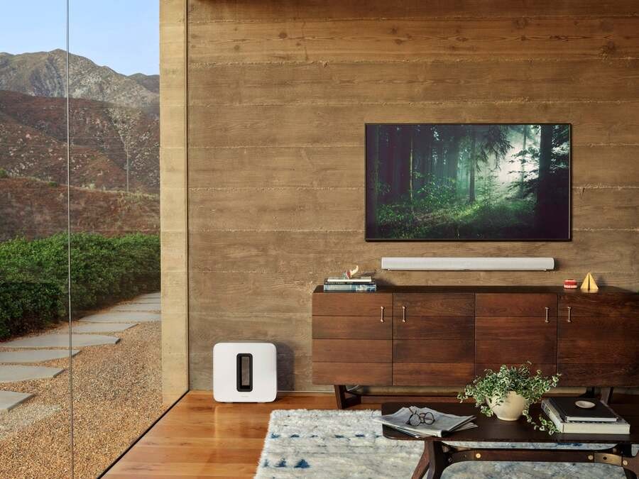 A luxury living room with high-end TV and a Sonos soundbar and subwoofer. The room oversees nature through a roof-to-ceiling window.