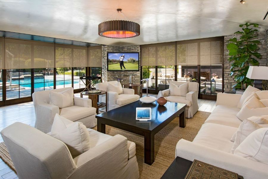 A living room with shades half drawn, a pool in the backyard, and a smart home system control on the coffee table. 