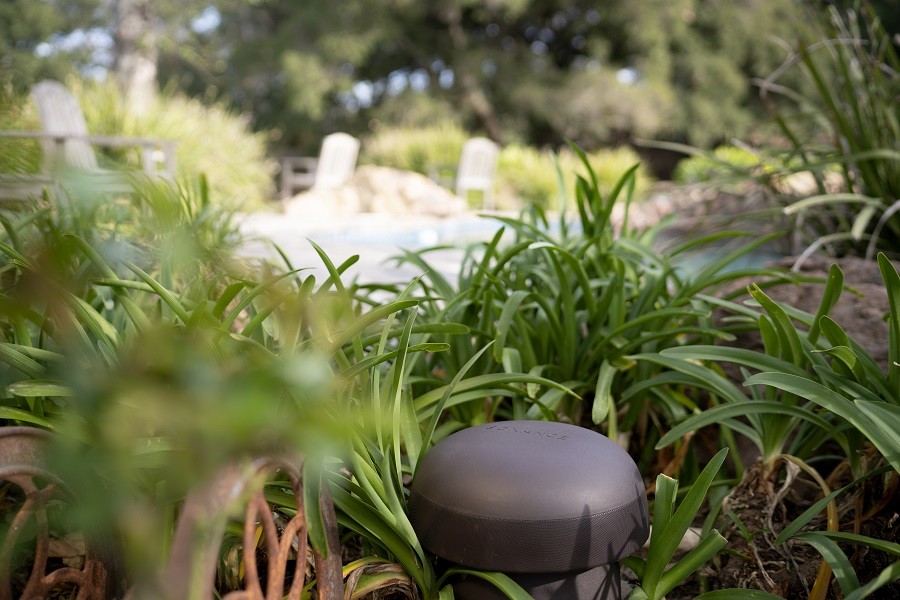 A speaker for outdoor entertainment placed in a garden bed with soft-focus patio furniture in the background. 