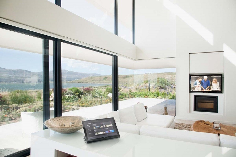 Sunlit minimalist modern living room with touchscreen and television looks over semi-wild garden and shoreline.