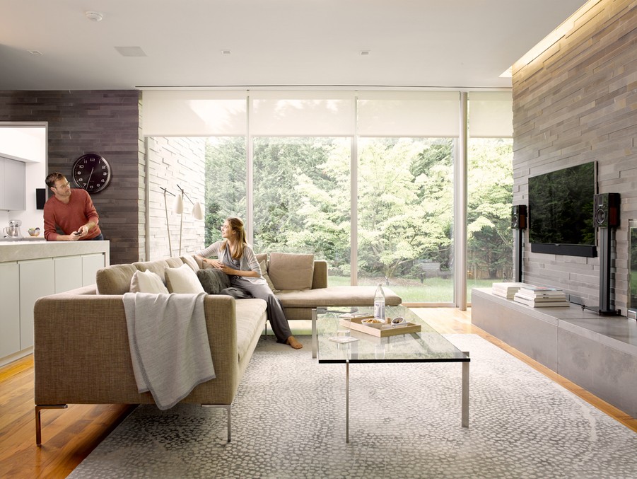 a couple in a living room with sofas, TV, glass table, and motorized shades.